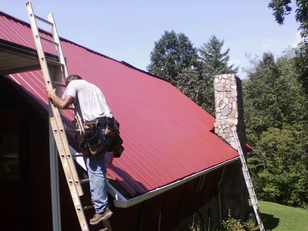 Roofer on a ladder beside a red metal roof and stone chimney — Soffit & Fascia Repair by Roofing Brothers in Chattanooga